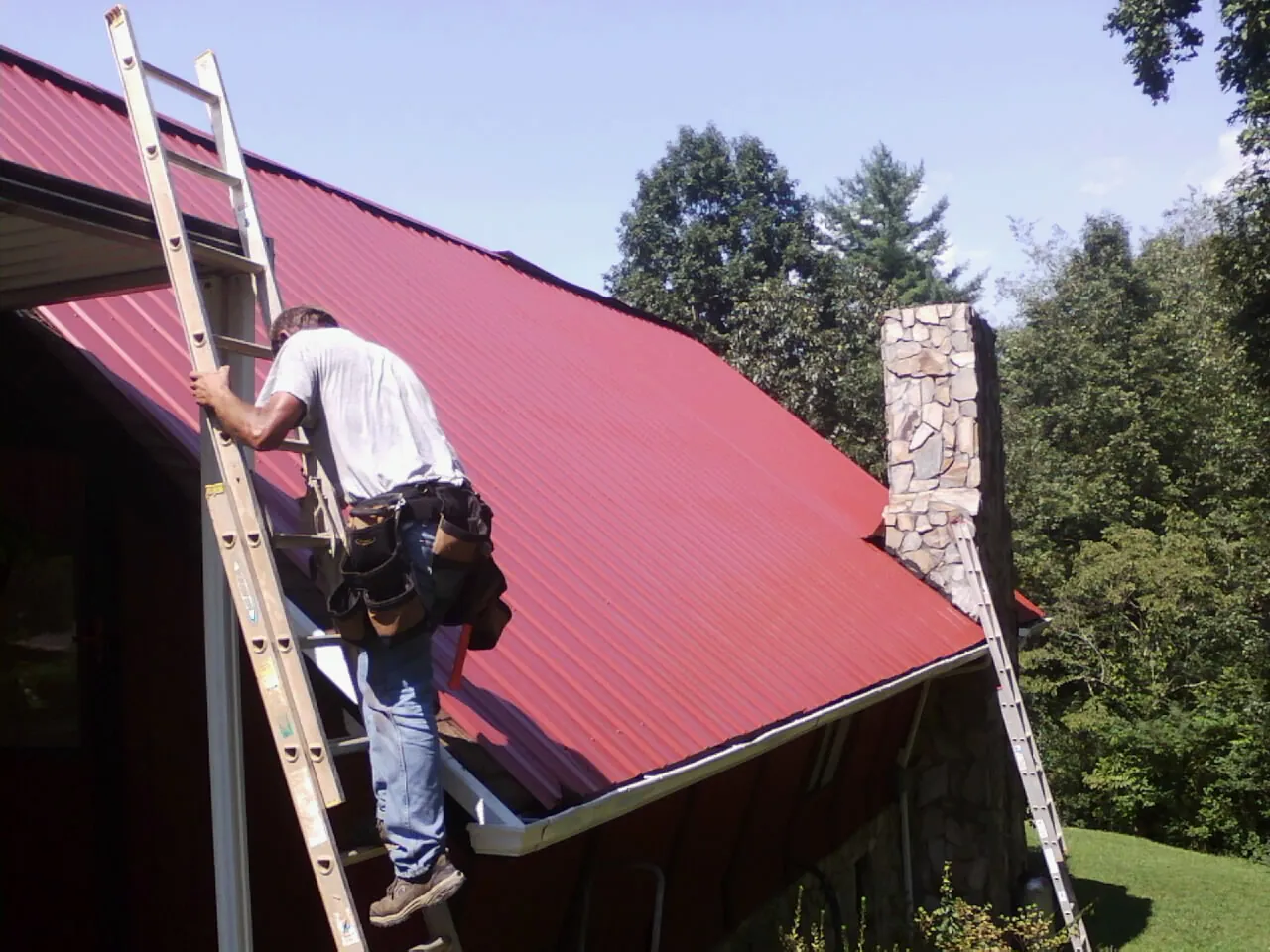 Roofer on a ladder beside a red metal roof and stone chimney — Soffit & Fascia Repair by Roofing Brothers in Olivehill